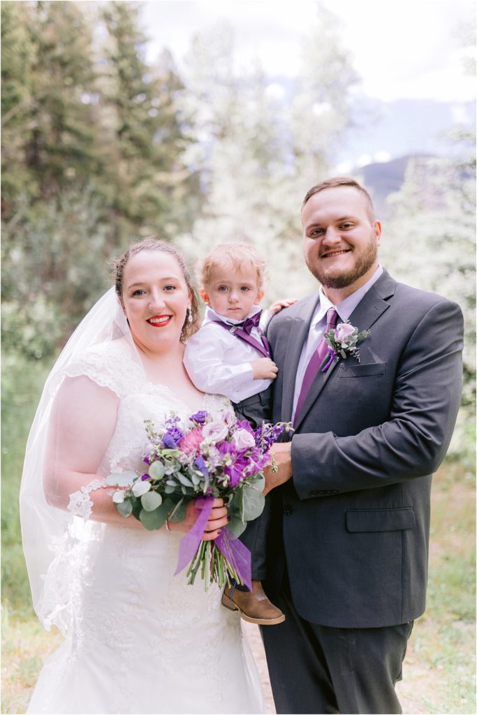 Bride and groom hold a small wedding guest in a tiny purple tuxedo, with a gorgeous purple floral arrangement too. Wedding was shot by local wedding photographer team Shayla Cristine Photography.