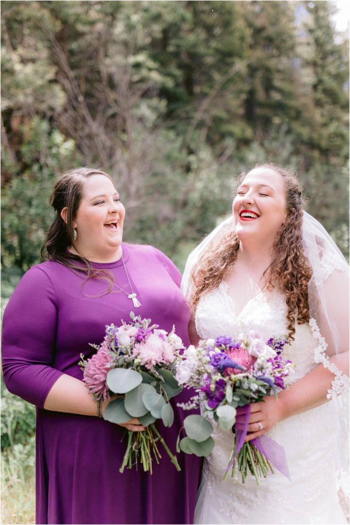 The bride and her bridesmaid laugh, holding wedding floral arrangements and in their bridesmaids gown and bridal gown - the perfect, happy moment captured by Shayla Cristine Photography.